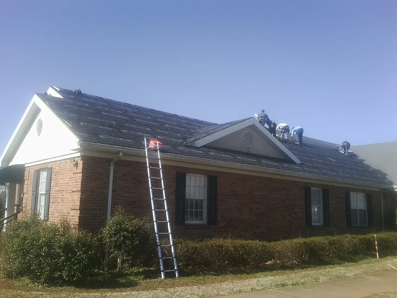 Crew tearing shingles off a brick ranch roof with a ladder set — Metal Roof Repair work by our crew in Island Lake