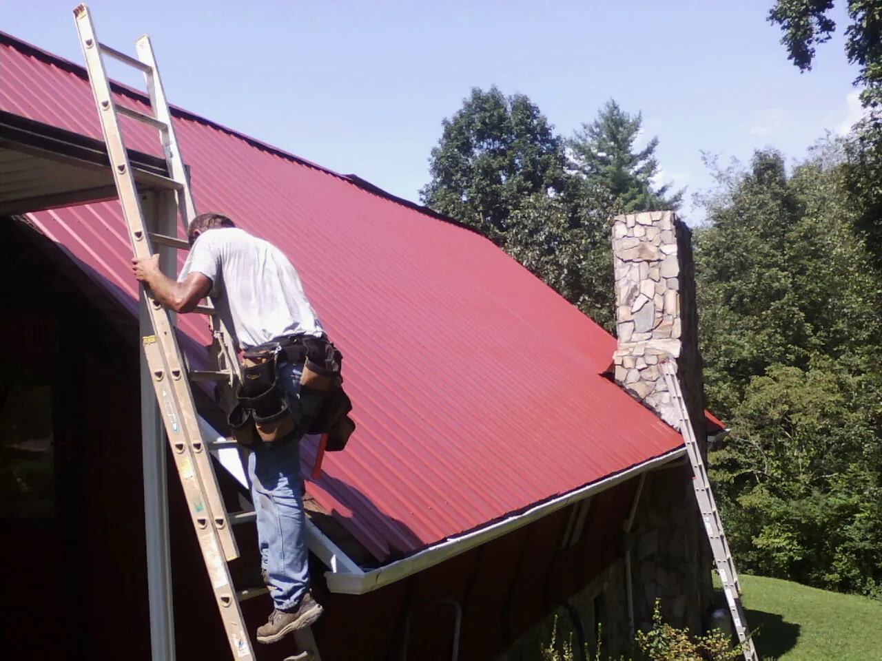 Roofer on a ladder beside a red metal roof and stone chimney — Soffit & Fascia Repair by Roofing Brothers in Island Lake